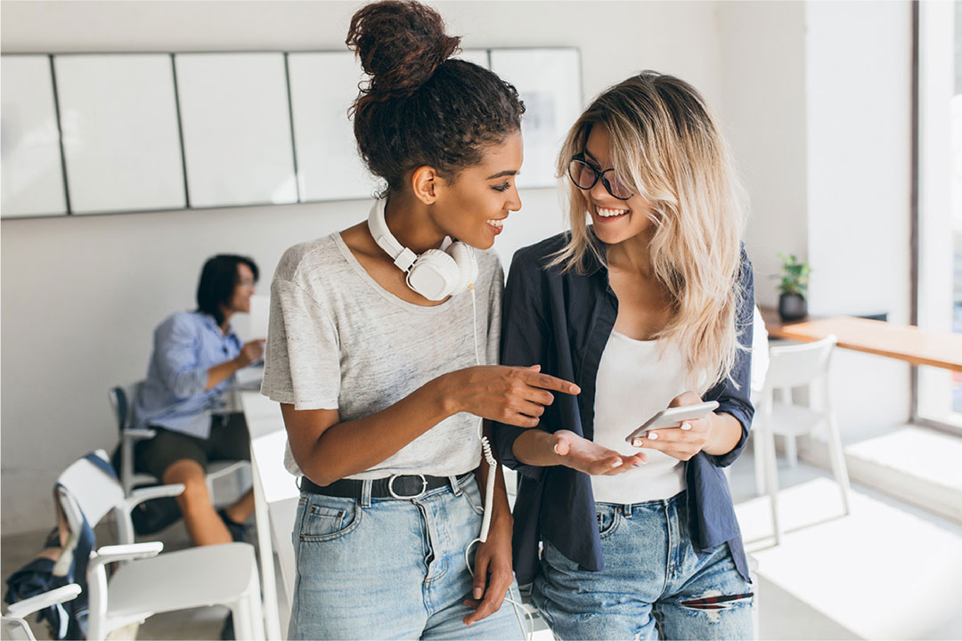 Due ragazze in un ufficio sorridono mentre guardano su uno smartphone la Web-App creata da YesWeWork per i propri dipendenti