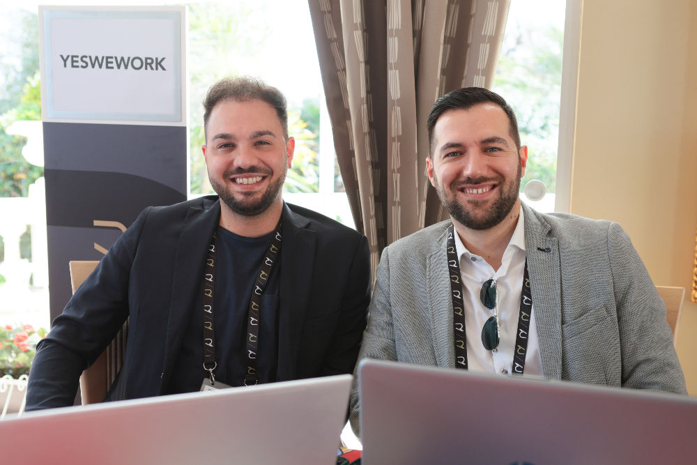 Antonio Porro e Antonio Giacobelli sorridono seduti dietro ai loro laptop allo stand di YesWeWork al Richmond HR Forum