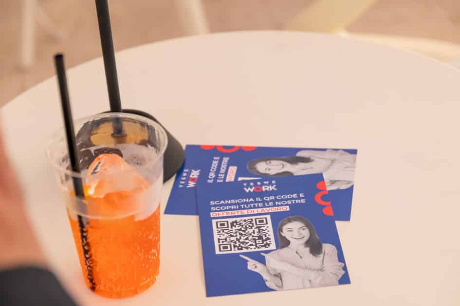 Su un tavolino dell'area aperitivo dell'ApeRecruiting Trani sono posati un bicchiere di spritz e dei volantini YesWeWork