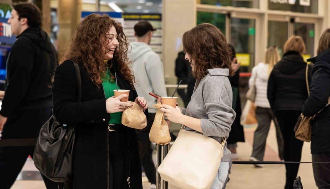Due ragazze chiacchierano e ridono durante l'ApeRecruiting; Hanno tra le mani un drink e un sacchetto di Finger Food