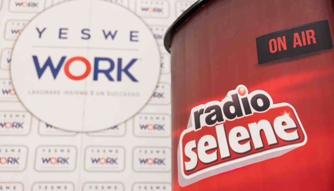 Inquadratura di un pannello brandizzato 'YesWeWork' dietro il banco regia di Radio Selene che riporta la scritta 'on air'