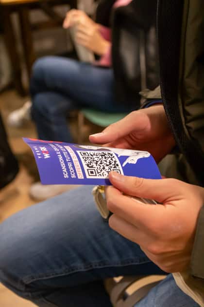 Le mani di un partecipante all'evento che osserva con attenzione il volantino di YesWeWork con un QR Code per cercare lavoro