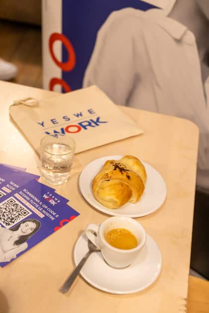 Su uno dei tavoli del Caffè Portineria sono poggiati una tazza di caffè, un croissant e una pochette con gadget di YesWeWork