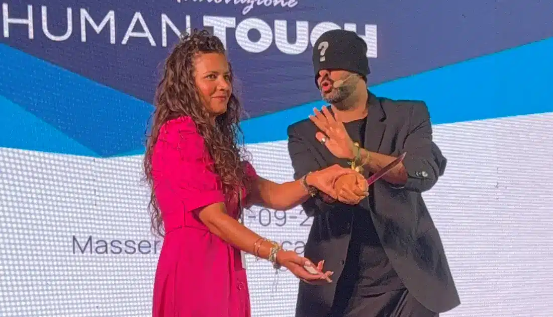 Andrea Paris, con il volto coperto da un cappuccio, interagisce con Santa del team YesWeWork sul palco durante un trucco