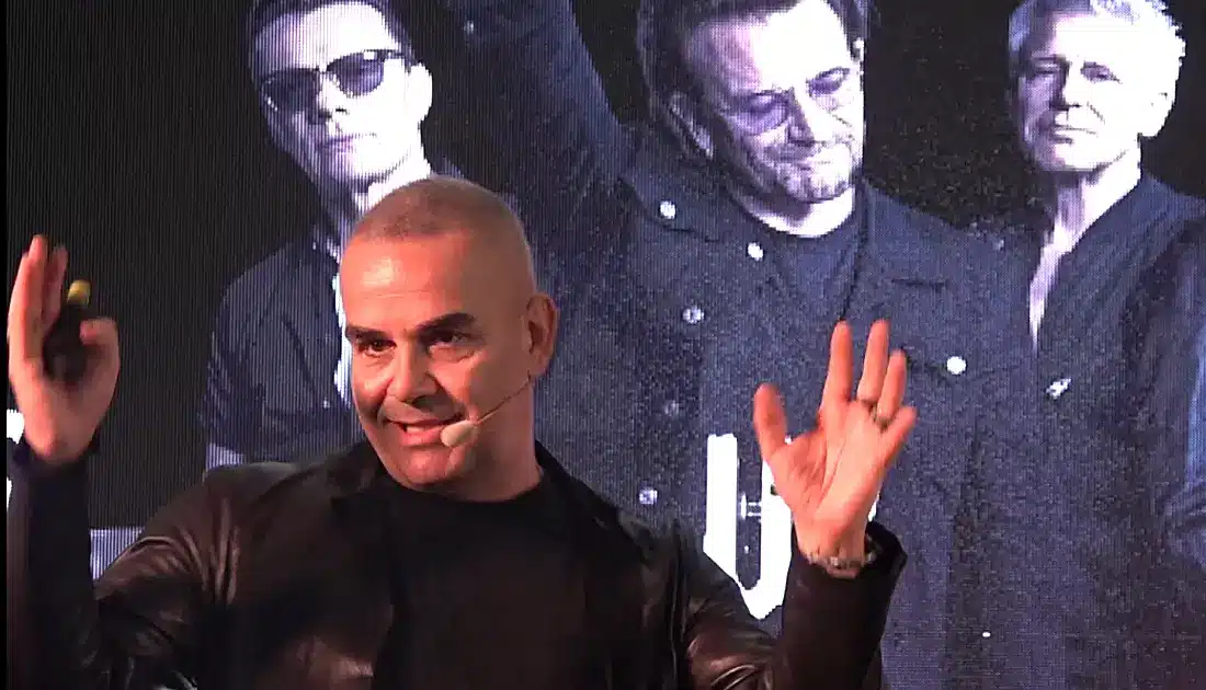 Primo piano di Max Panìco durante il suo intervento. Sul maxischermo alle sue spalle si intravede una fote degli U2