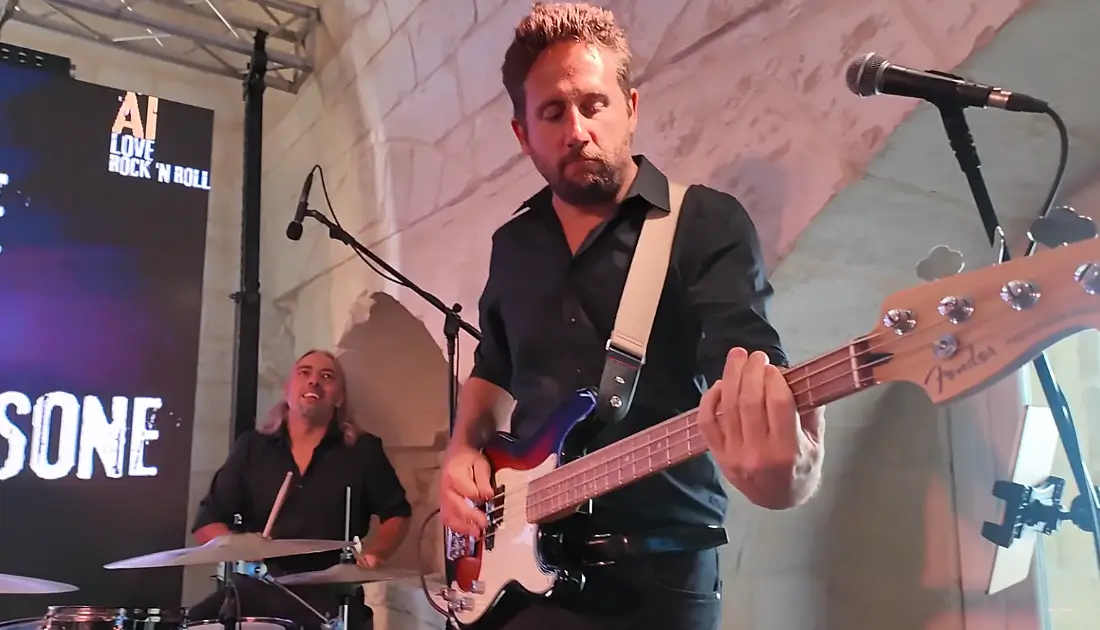 Inquadratura ravvicinata del bassista della band di 'Rock in Azienda'; alle sue spalle si intravede il batterista